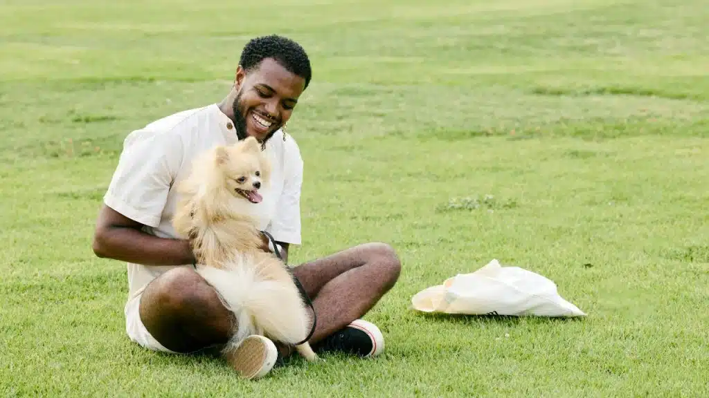 A smiling man sits on the grass and holds a small fluffy dog.