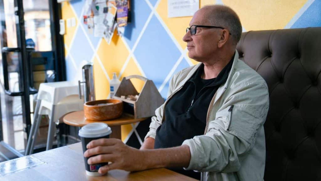 A man with glasses holds a paper coffee cup while sitting at a cafe table.