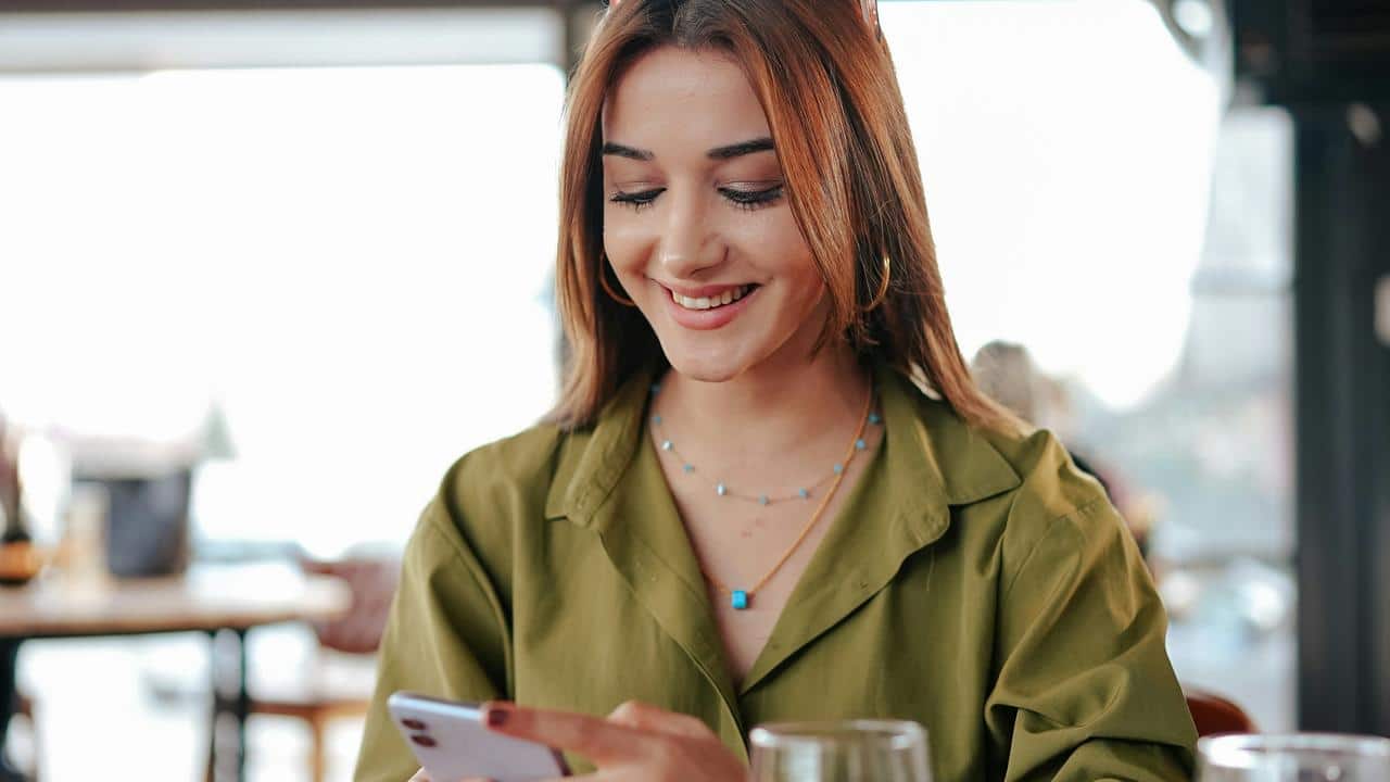 A smiling woman in a green shirt looking at her smartphone in a café.