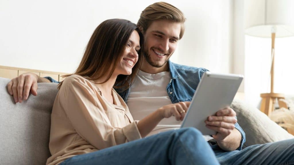 A smiling man and woman sit on a couch together looking at a white tablet.