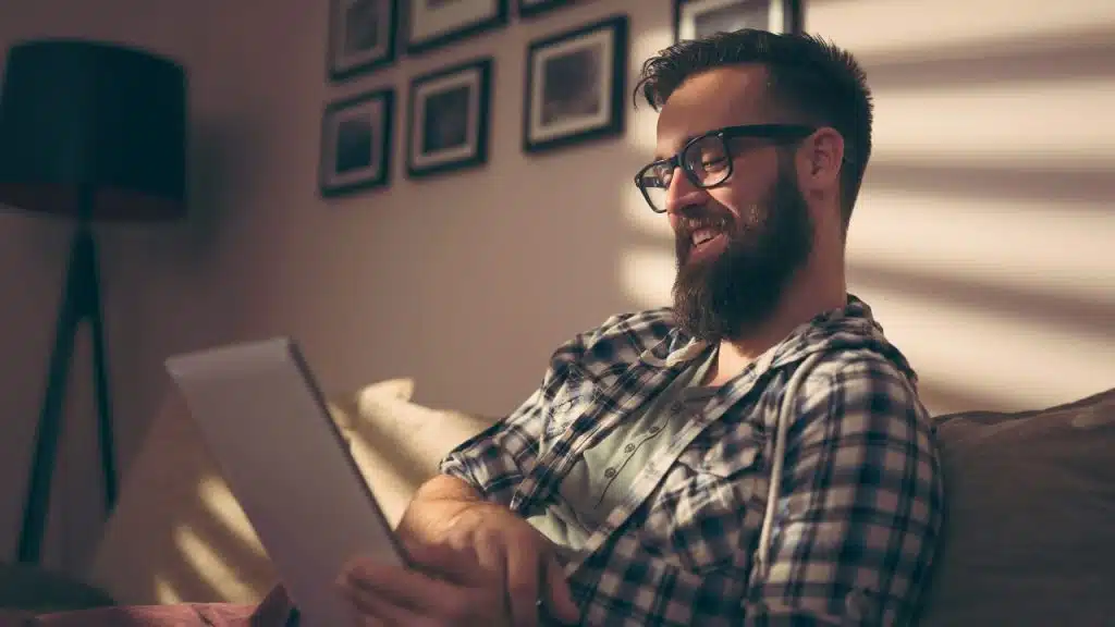A man with a beard and glasses smiles while looking at a digital tablet indoors.