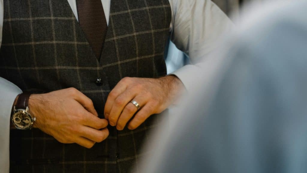 A man in a vest and tie adjusts his clothes, showing a wristwatch.