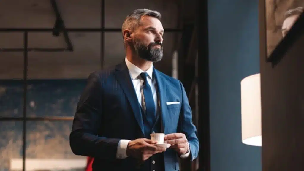 A bearded man in a suit holding a small cup standing in an office.