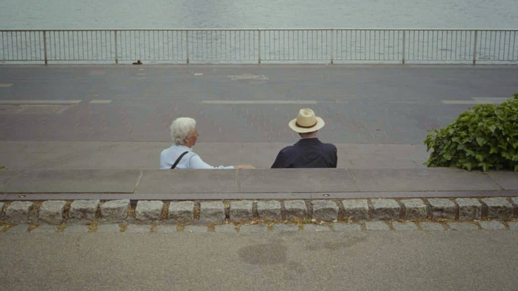 A couple sitting apart from each other on a bench by the street along the sea.