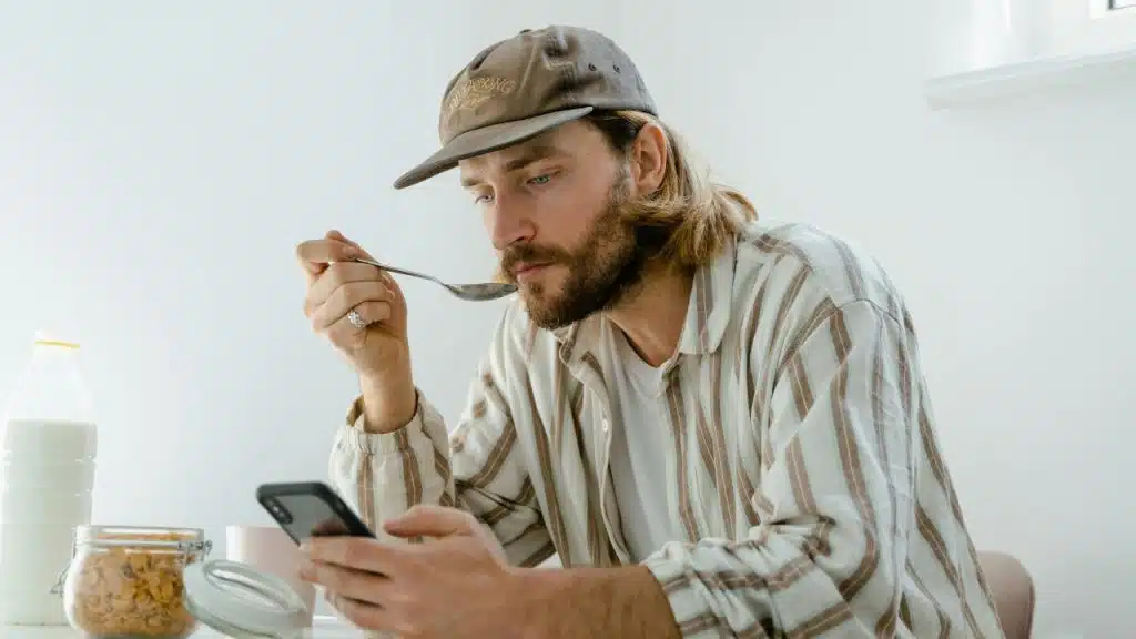 A man with a beard and baseball cap eats while looking at a mobile phone.