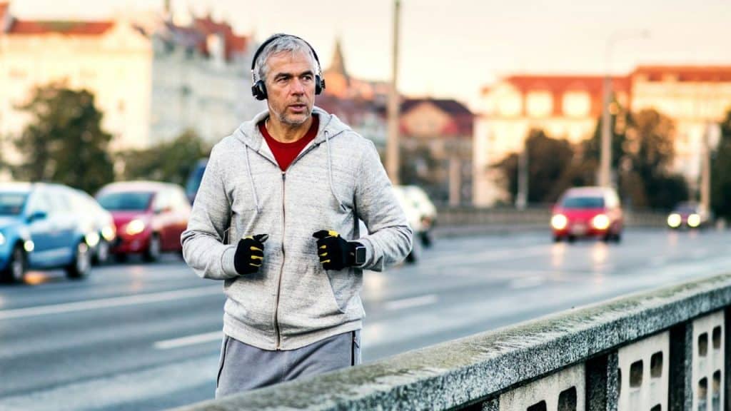 A man with headphones jogs across a bridge with city traffic and buildings in the background.