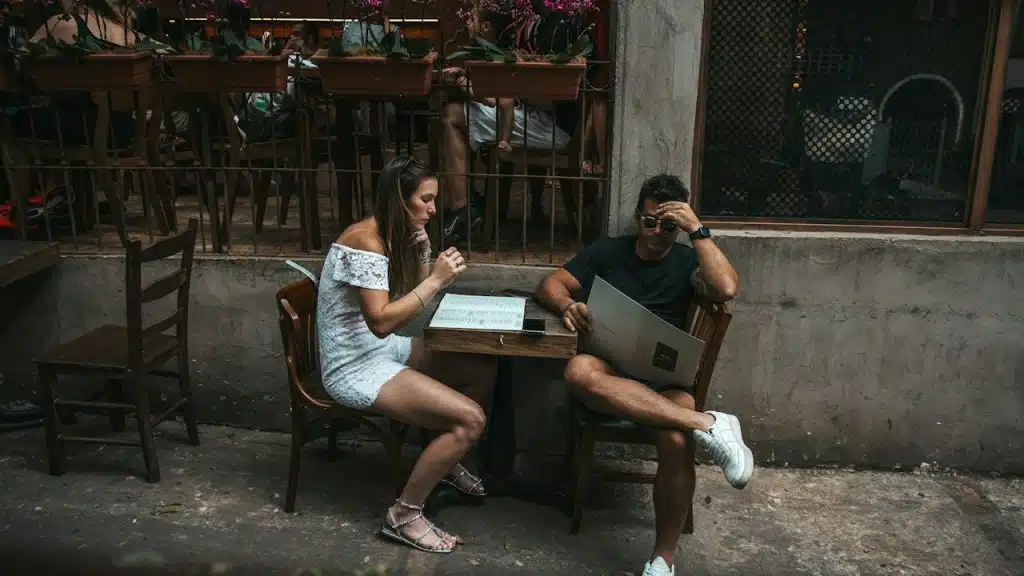 A woman in a white off-shoulder dress and a man holding a laptop sitting at a table in an outdoor cafe