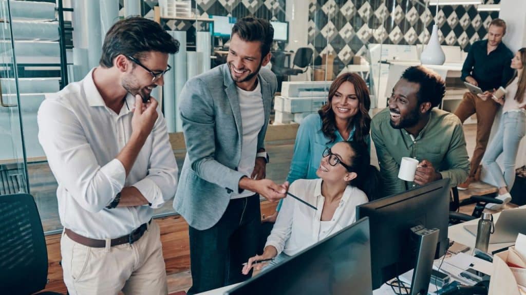 A diverse group of colleagues smile and talk while gathered around an office computer.