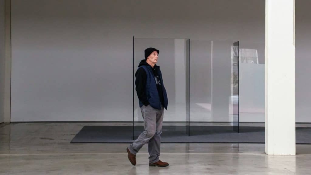 A man wearing a hat and vest walks across a large, empty room.