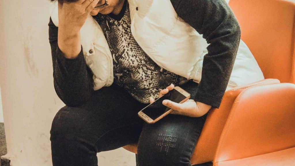A worried person sitting on a chair holding a smartphone and covering their face