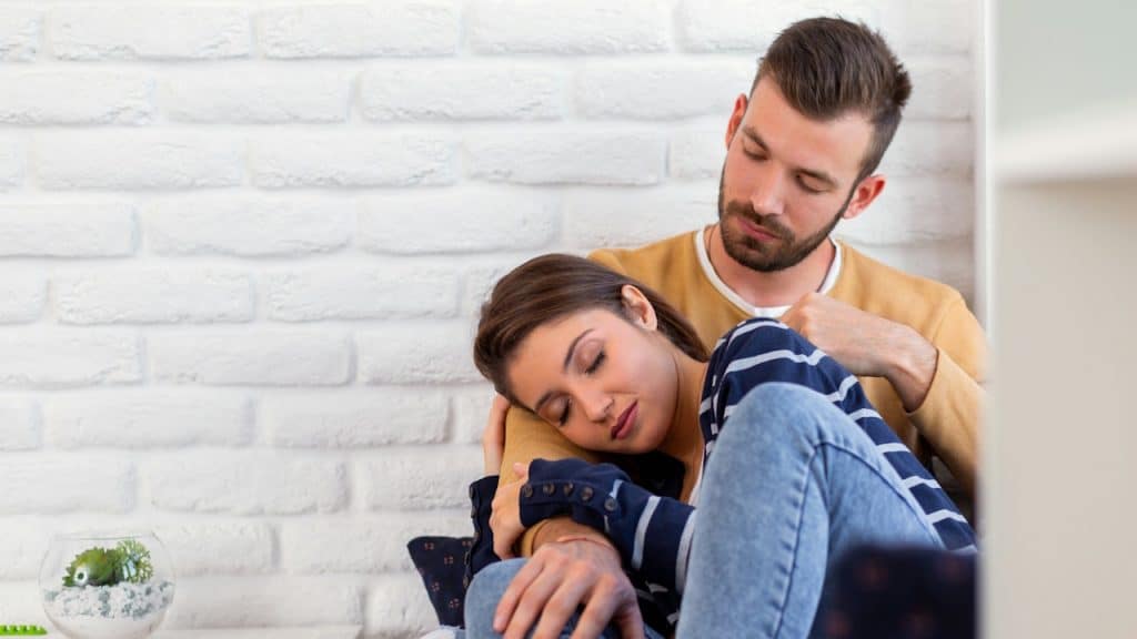 Young man comforting a woman who is leaning her head on his shoulder in a domestic setting with a white brick wall background.
