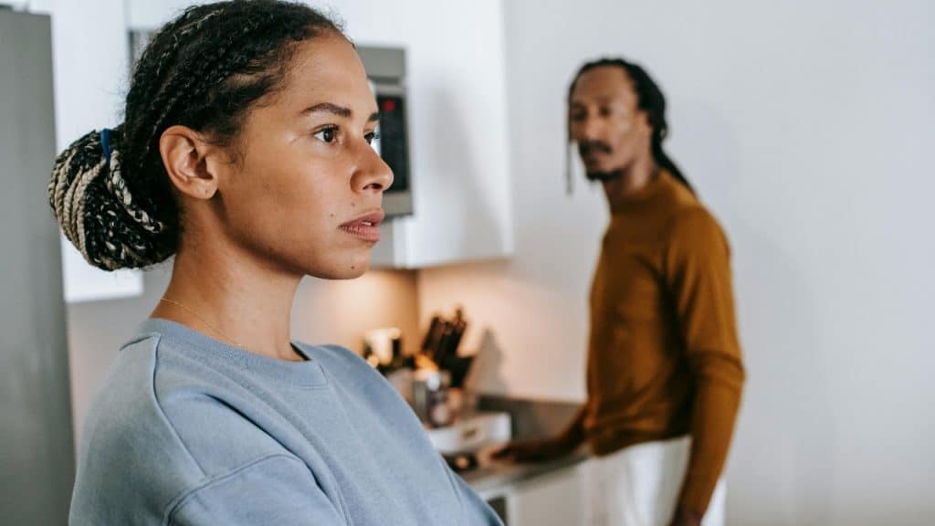 A woman looks away with a serious expression while a man stands blurred behind her.