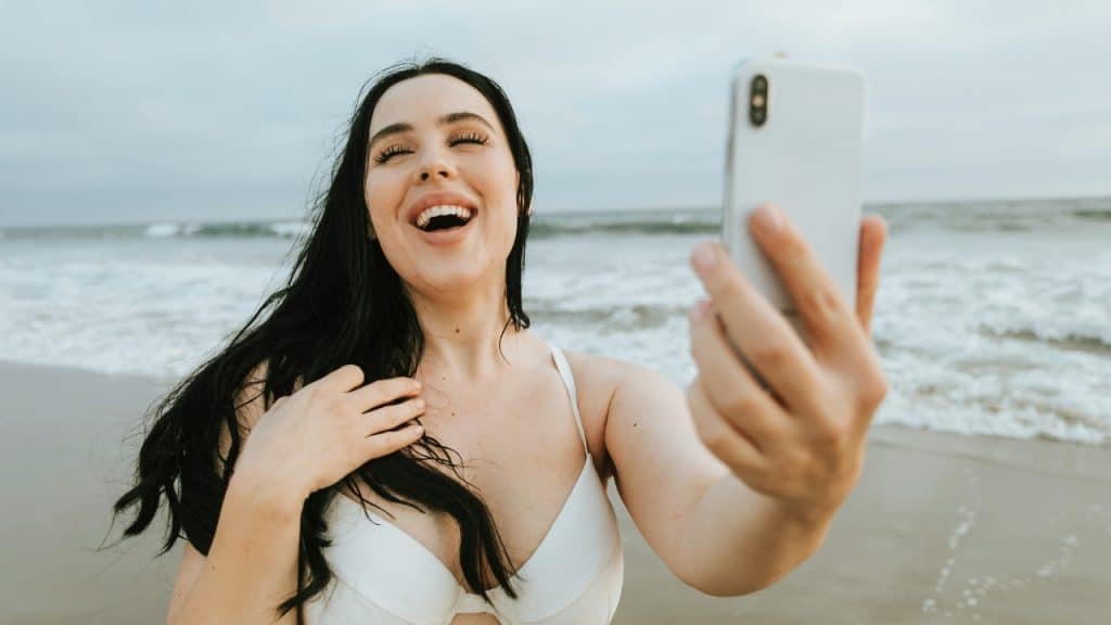 A woman in a white bikini laughs while holding up a phone to take a selfie.