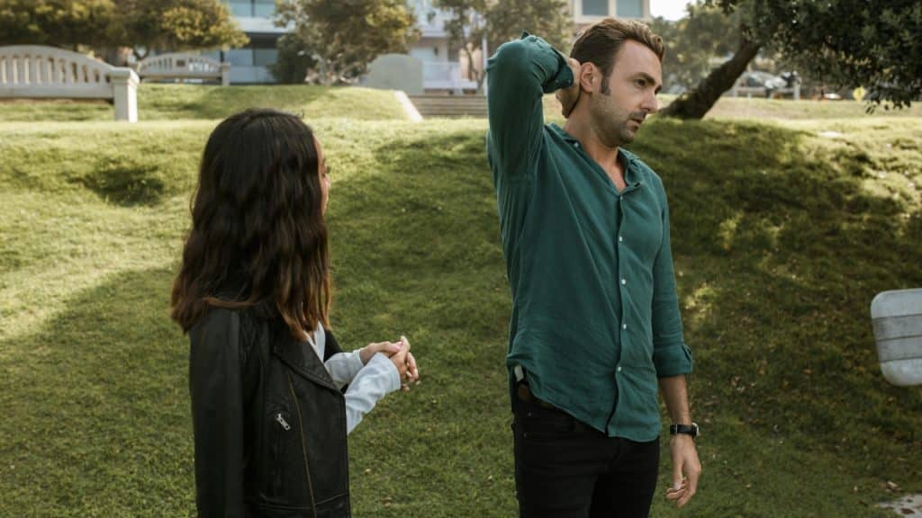 A woman in a leather jacket stands near a man holding his neck outdoors.