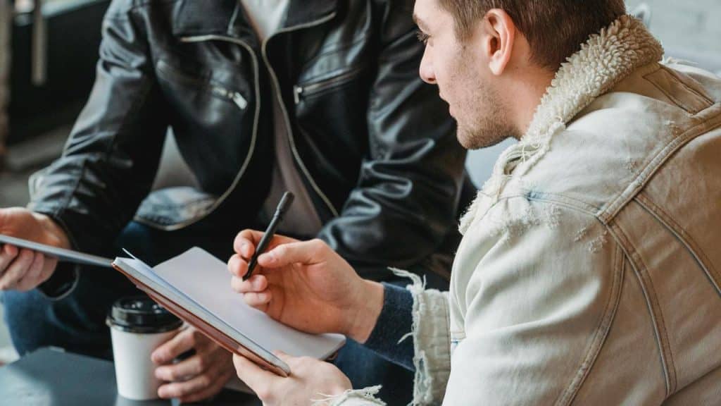 A man writing in a notebook while sitting and talking with another man holding a tablet.