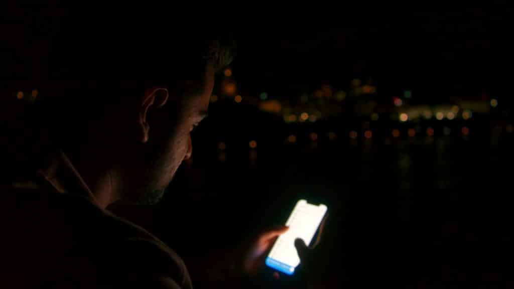 Silhouette of a man looking at a glowing smartphone screen against a dark nighttime background.