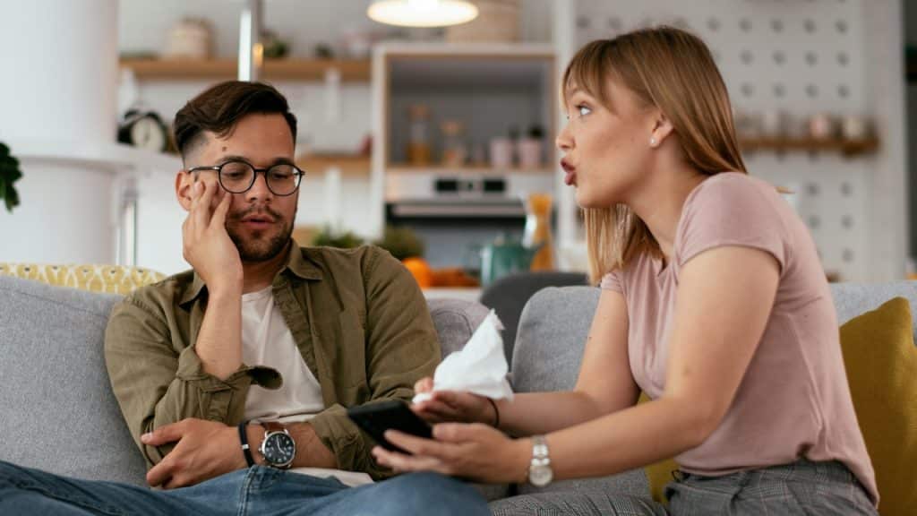 A woman holds a tissue and speaks to a man sitting on a couch.
