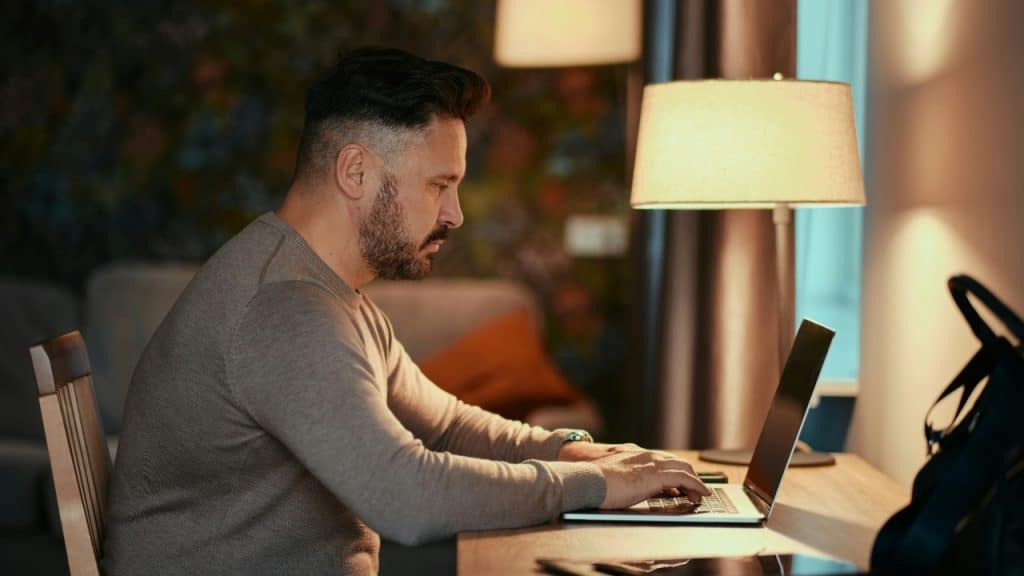 A man in a sweater works on a laptop at a desk at night.