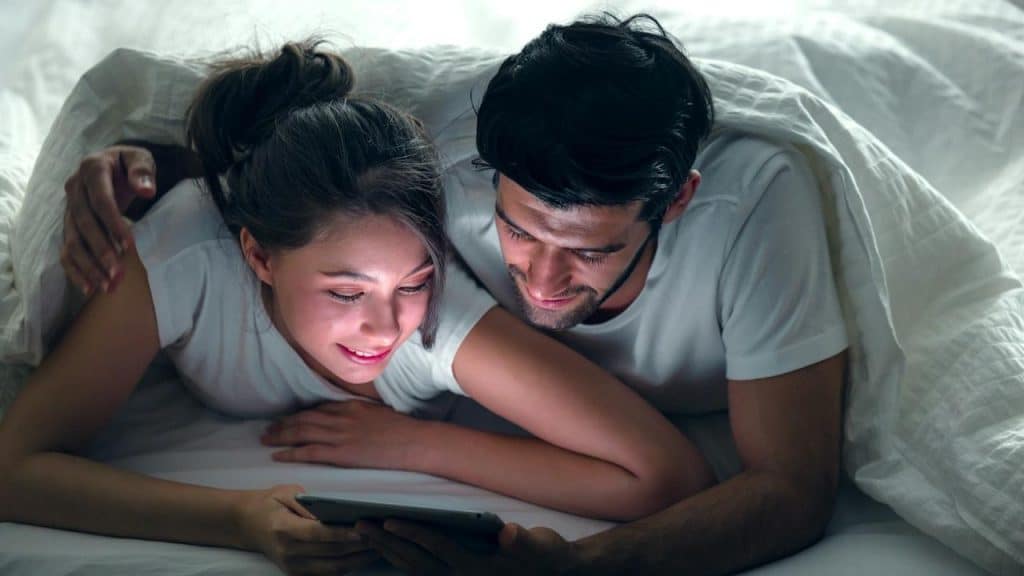 A young, sweet couple on a bed watching something on a tablet gadget and relaxing in the bedroom.