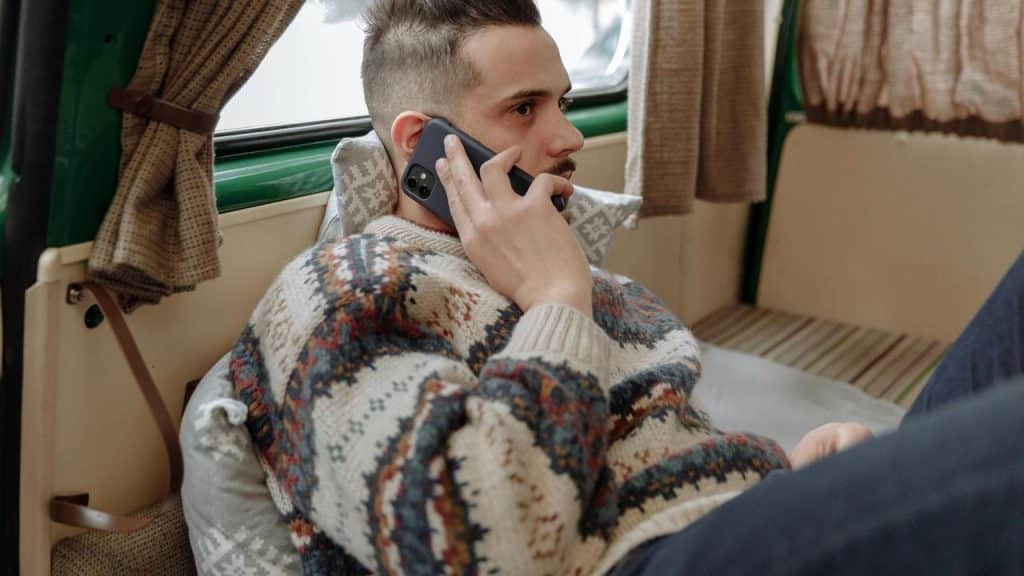 A man in a patterned sweater talking on a smartphone while sitting inside a camper.