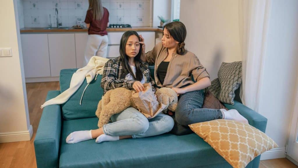 A woman comforts a sad teen on a couch while another person stands in the kitchen