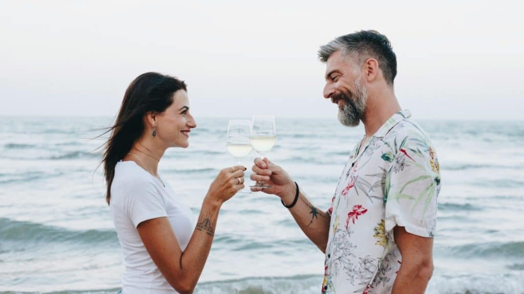 A man and woman smile at each other while clinking wine glasses on a beach.