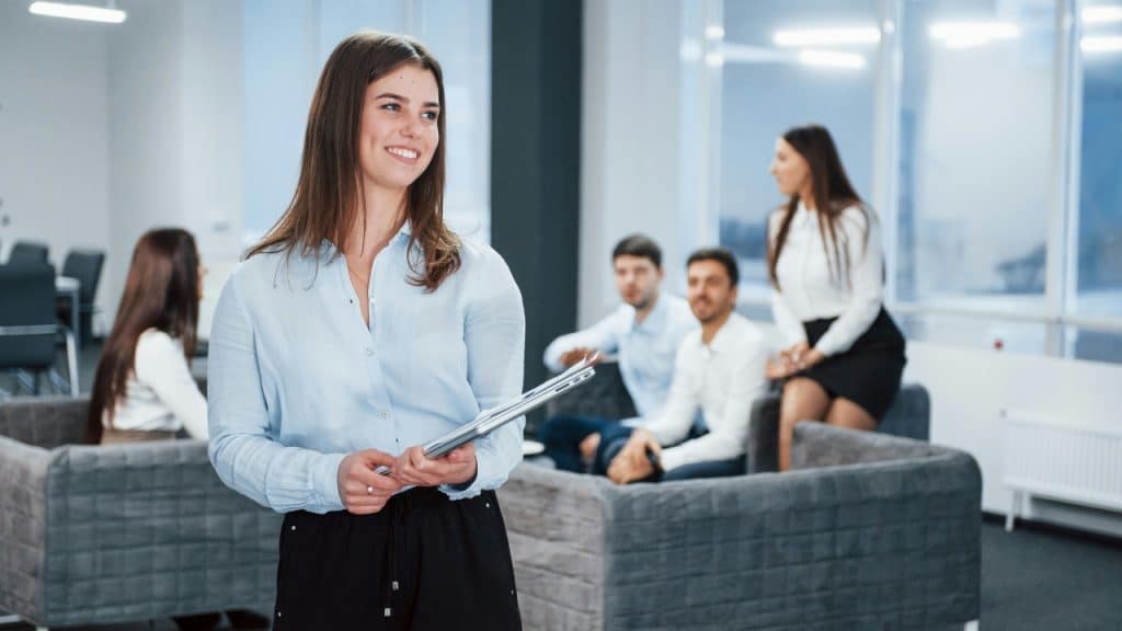 A smiling woman in a light blue shirt holds a laptop in a bright office.