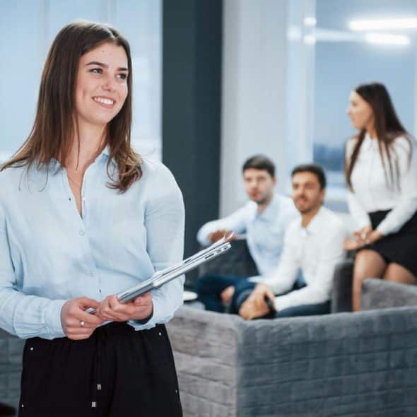 A smiling woman in a light blue shirt holds a laptop in a bright office.