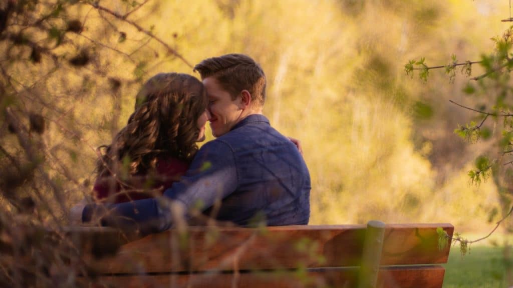 A couple sitting together on an outdoor wooden bench, looking closely at each other.