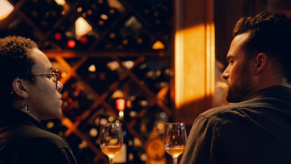 Two people sit at a table with wine glasses in a dimly lit setting.
