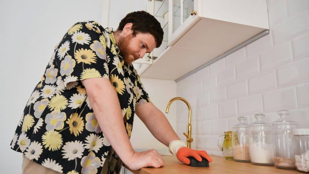 A man in a floral shirt wearing gloves wipes a kitchen counter surface.