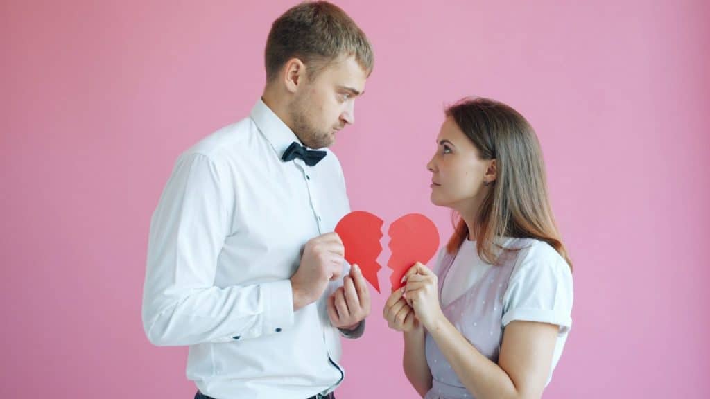 A man and a woman facing each other while holding two halves of a broken red heart against a pink background