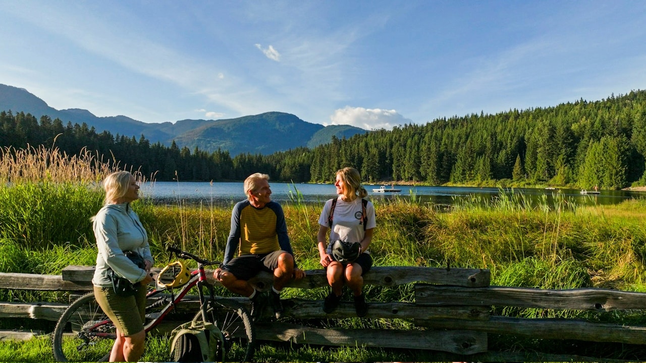 Family with mountain bikes pauses against fence by lakeshore at sunset in Whistler.