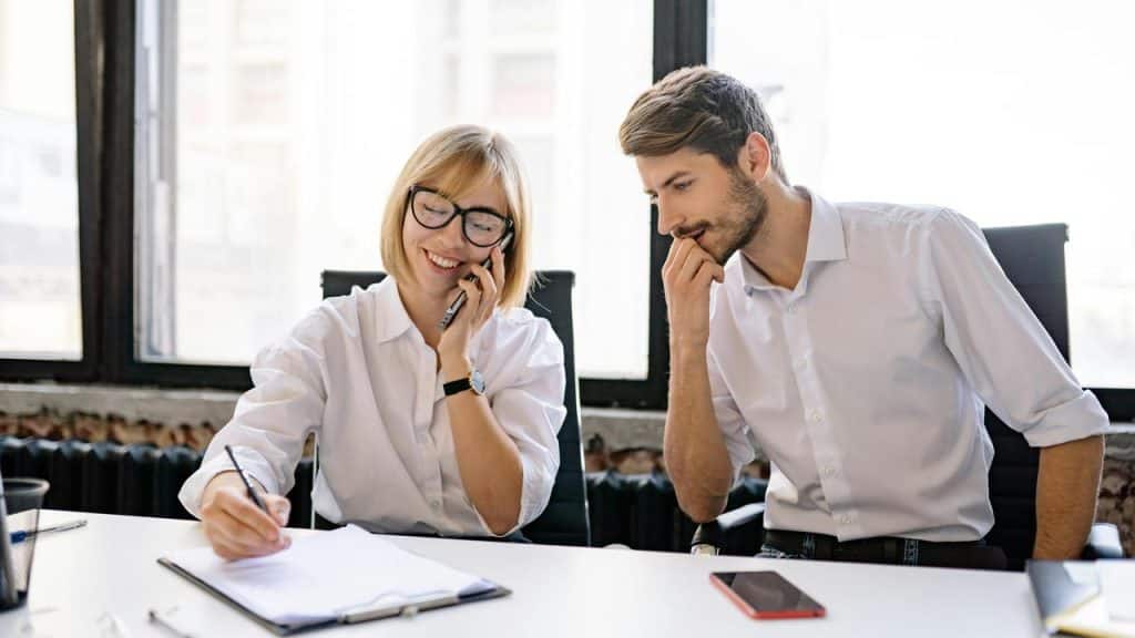 A woman on the phone writing notes while a man watches