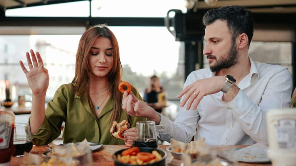A man and woman sit at a restaurant table with food, gesturing during a conversation.