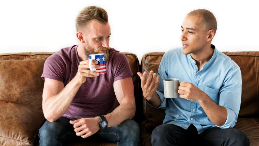 Two men sit on a leather sofa holding mugs while one man gestures during conversation.