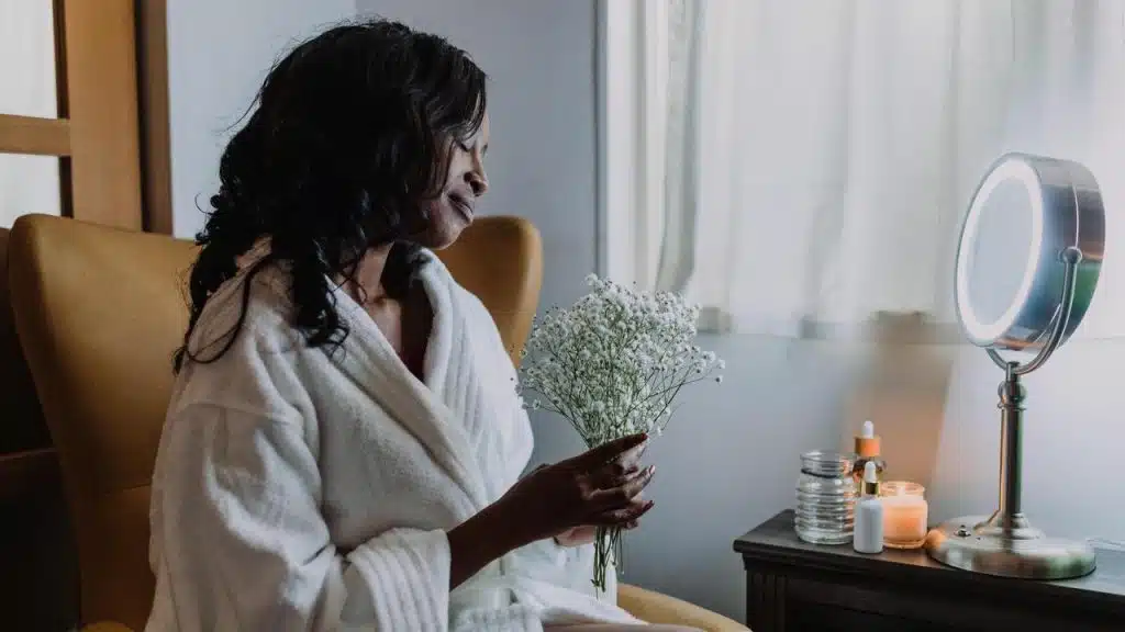 A woman in a robe holding a small bouquet of flowers while sitting beside a lit vanity mirror.