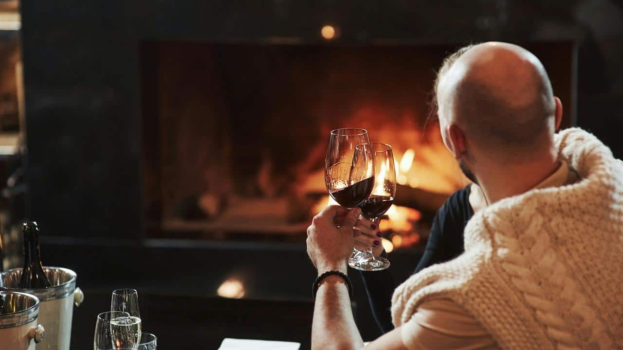 A couple clinks wine glasses by a fireplace.
