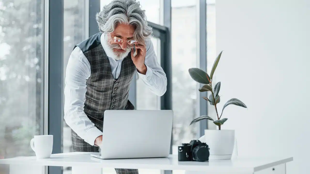 An older man wearing glasses is using his laptop while talking on the phone.