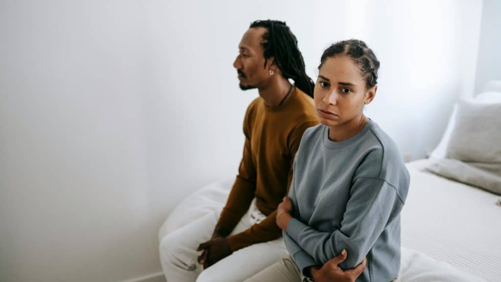 A man and woman sit on the edge of a bed facing different directions.
