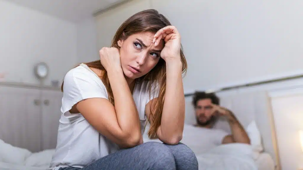 A woman looks away with a concerned expression while a man sits blurred behind her.