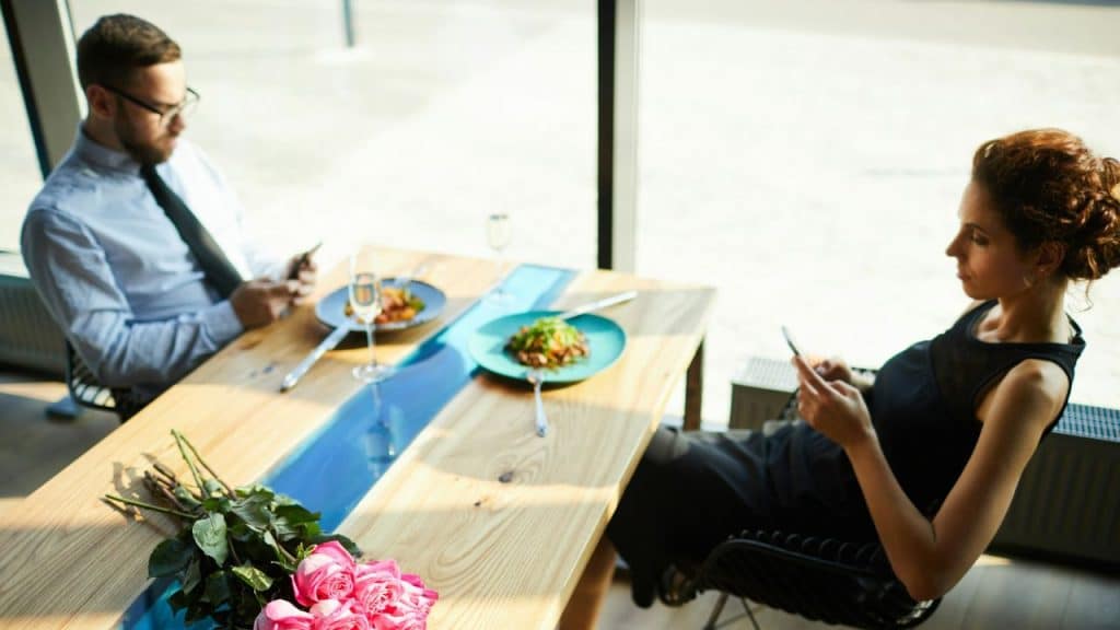A man and woman sit at a table with food, both looking at their smartphones.
