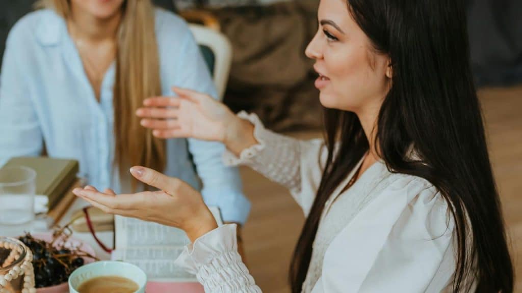 A woman gesturing while talking to another person at a table with coffee and books.