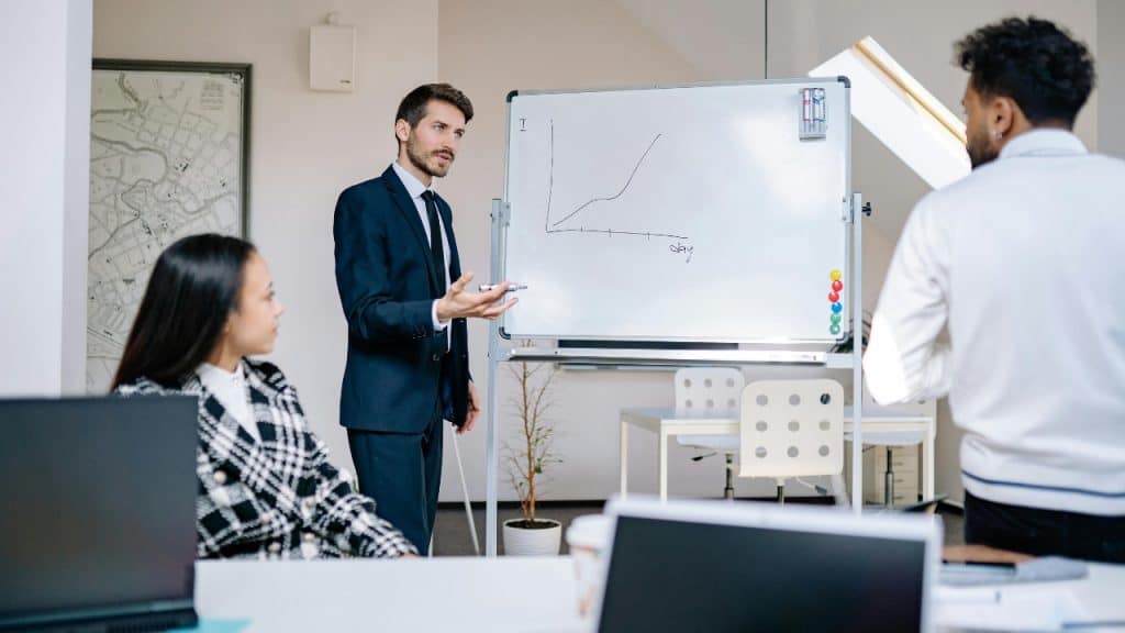 A man in a suit gestures toward a whiteboard with a line graph during a meeting.