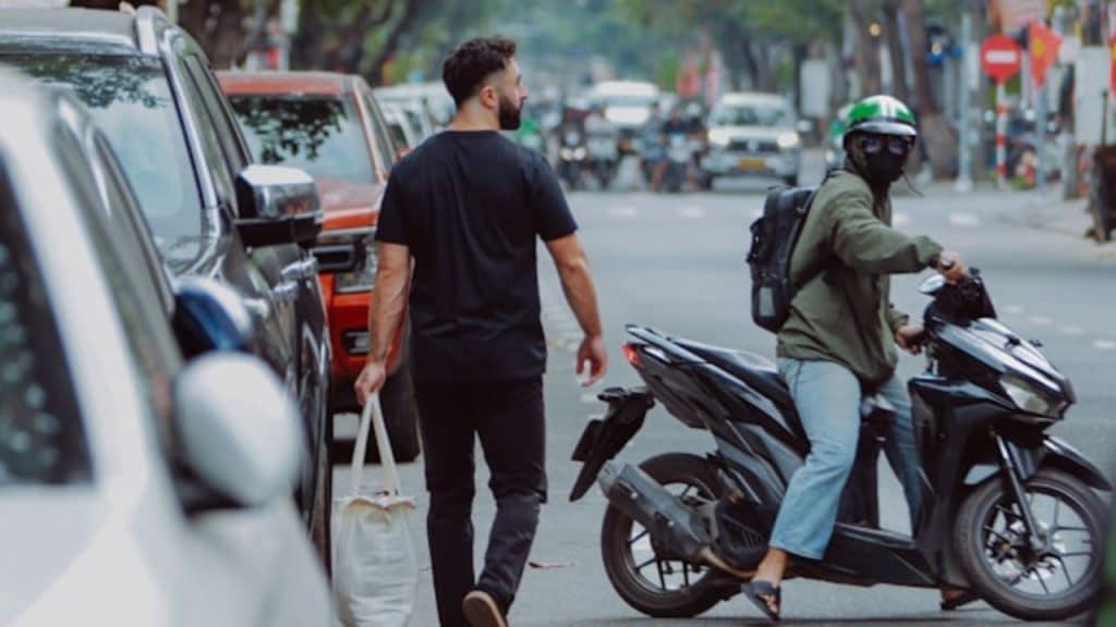 A man carrying a white bag walks past a person sitting on a motorbike.