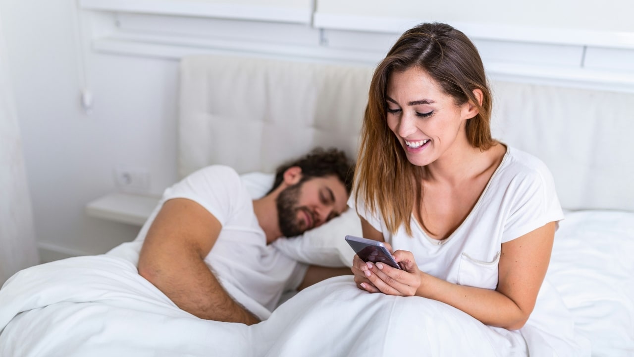 A smiling woman looks at her phone while a man sleeps in a bed behind her.