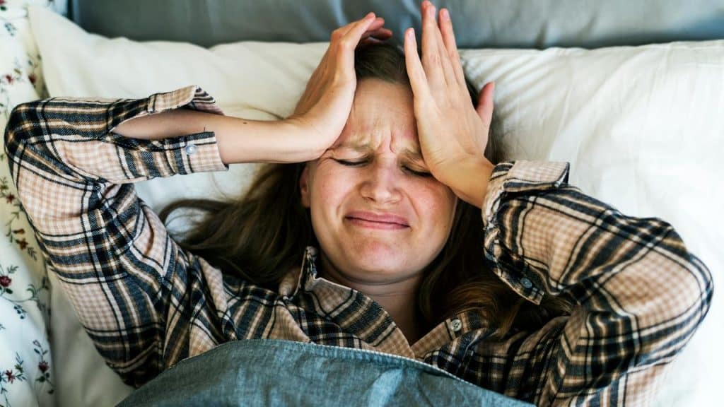 A woman lying in bed holding her head and looking distressed.