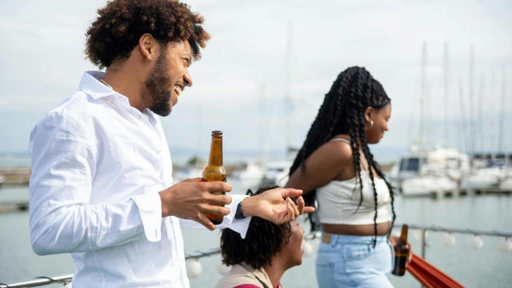 A group of friends laughing and talking while holding beer bottles on a boat near a marina.