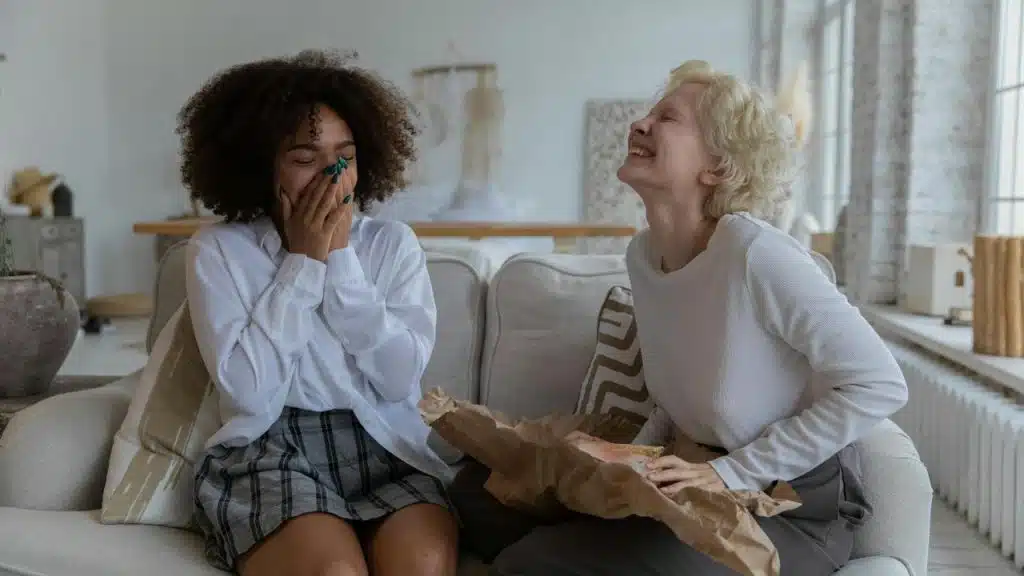 A pair of women laughing on a couch, one holding a wrapped gift.