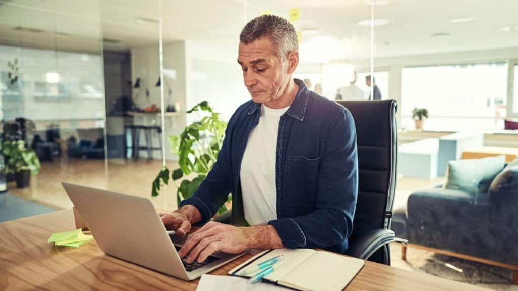 A man in a denim shirt types on a laptop at a wooden desk in an office.